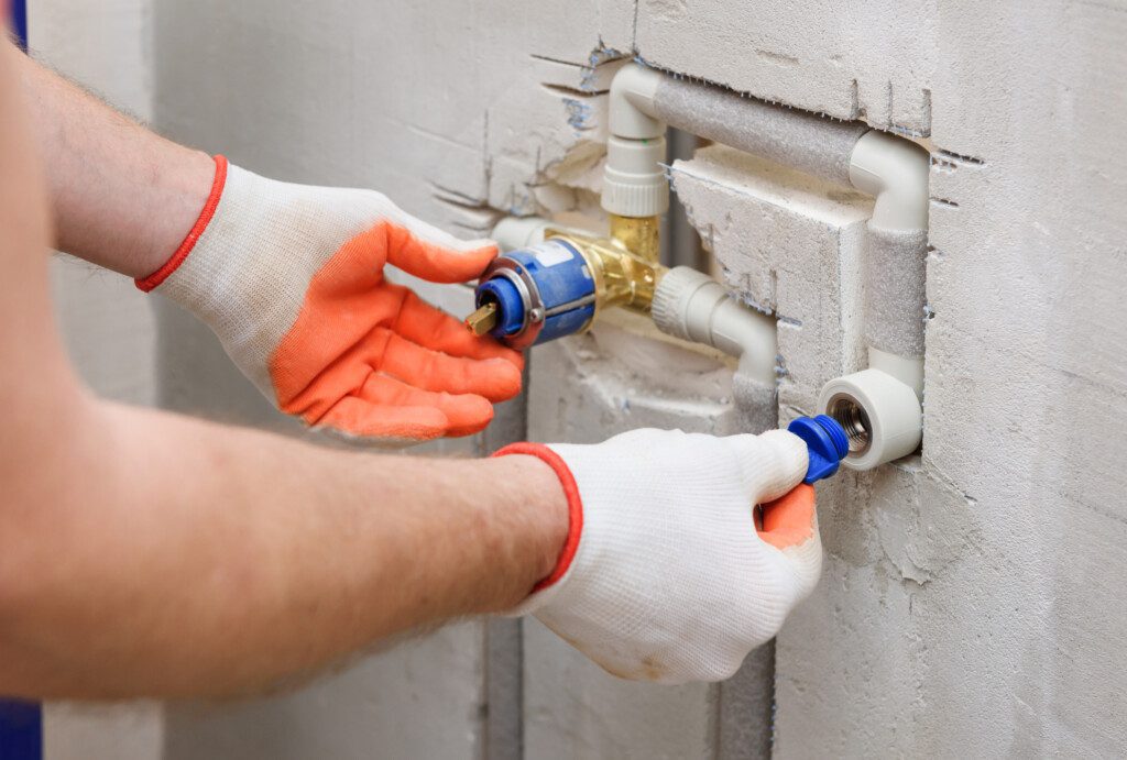 Imagem de um homem com usando luvas nas cores laranja e branco nas mãos consertando um registro de chuveiro instalado na rede hidráulica de uma parede branca