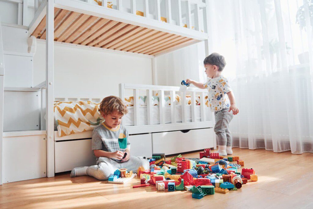 Dois irmãos brincam em seu quarto com alguns brinquedos no chão.