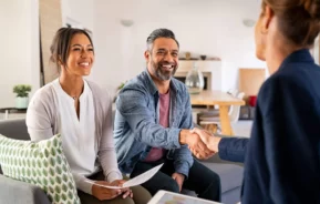 Imagem de um casal formado por um homem e uma mulher sentados no sofá de um imóvel enquanto o homem aperta a mão de uma agente imobiliária em sinal de alegria para ilustrar matéria sobre o repasse de financiamento