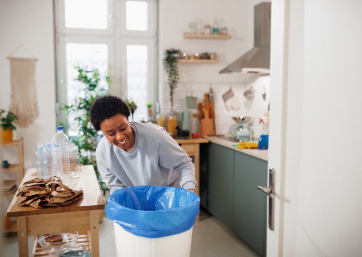Imagem de uma mulher colocando um saco de lixo azul em uma lixeira branca na cozinha de casa