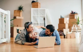 Imagem de uma família feliz de três pessoas descansando no chão de um imóvel, sorrindo e assistindo a algo em um laptop, cercados por caixas desempacotadas de uma mudança.