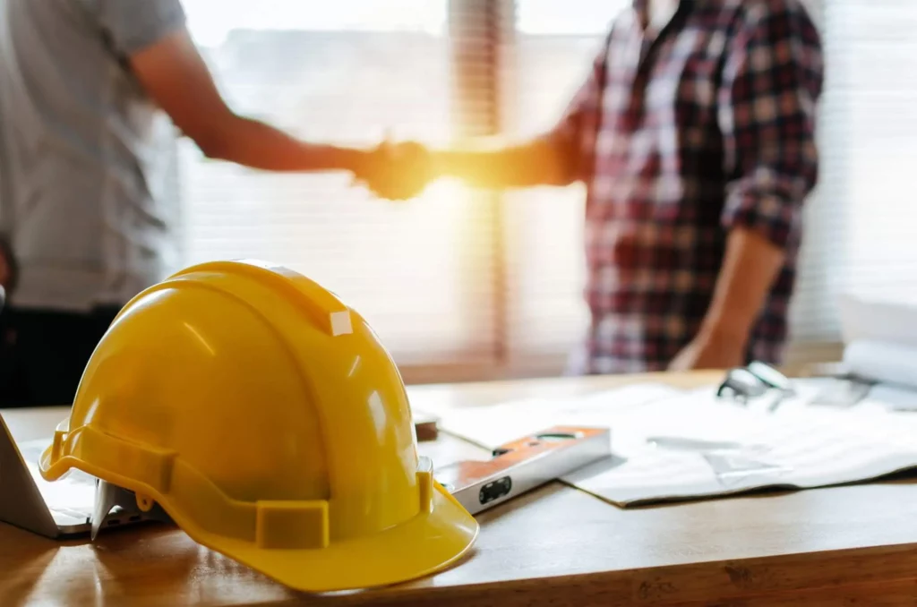 Imagem de um capacete de trabalho de construção civil em cima de uma mesa em primeiro plano enquanto um homem aperta a mão de um engenheiro em segundo plano para ilustrar matéria sobre como tirar habite-se de imóvel já construído