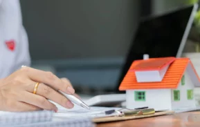 Imagem de um agente imobiliário analisando documentos em cima de uma mesa onde também estão um computador, algumas cadernetas e uma casa de madeira em tamanho miniatura para ilustrar matéria sobre o habite-se