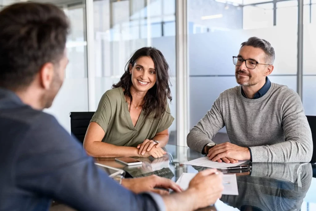 Imagem de um casal formado por um homem e uma mulher sentado em uma mesa em frente a um agente imobiliário enquanto seguram alguns papéis com as mãos para ilustrar matéria sobre o que é alienação fiduciária