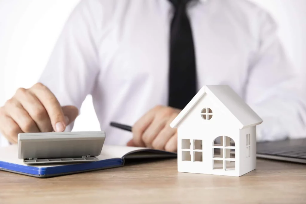 Imagem de um homem fazendo contas em uma calculadora que está ao lado de uma casa de madeira em tamanho miniatura para ilustrar matéria sobre quanto tempo demora o habite-se