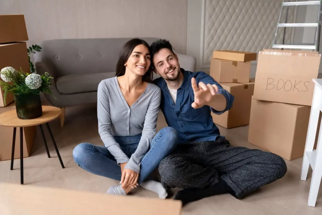 Imagem de um casal formado por um homem e uma mulher sentados no chão da sala de um imóvel em frente ao sofá e do lado de algumas caixas de papelão para ilustrar matéria sobre o significado de alienação fiduciária