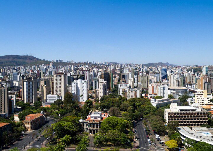 Foto que ilustra matéria sobre as regionais de Belo Horizonte mostra uma vista do alto da cidade, com destaque, me primeiro plano, para árvores da Praça da Liberdade. Ao fundo, prédios e mais atrás a Serra do Curral e um céu azul em um dia claro.