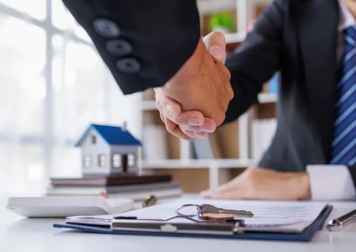 Dois homens apertam as mãos fechando um contrato. Há uma chave em cima do contrato e a miniatura de uma casa.