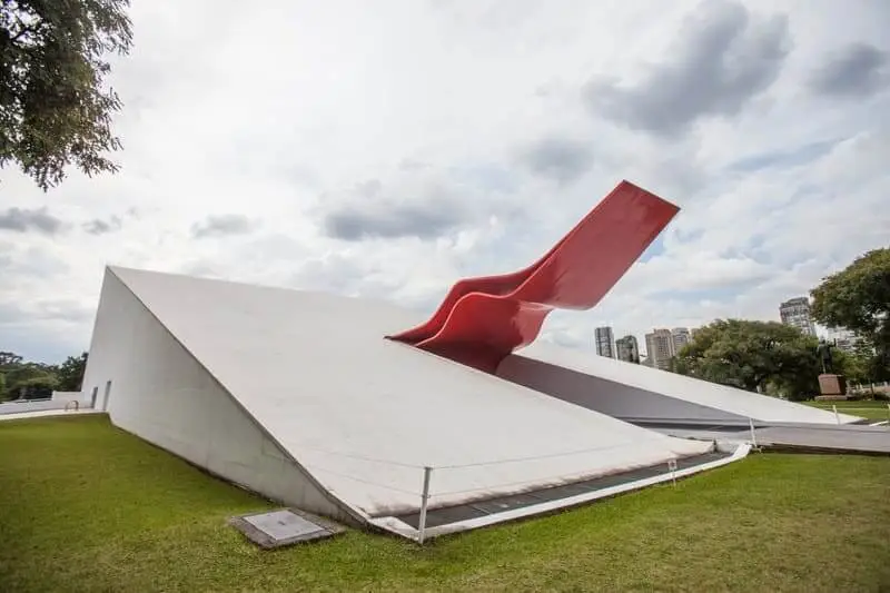 magem do Auditório Ibirapuera, prédio projetado por Oscar Niemeyer, mostra estrutura branca com paredes inclinadas e uma ousada estrutura vermelha emergindo do centro em meio à grama verde e às árvores. Prédios da cidade se erguem em um fundo nublado.