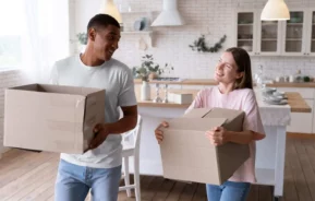 Imagem de um casal composto por um homem e uma mulher carregando caixas de papelão dentro de uma casa para ilustrar matéria sobre as cidades do interior de SP