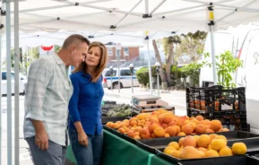 Imagem de um casal sorridente formado por um homem e uma mulher em frente a uma banca de frutas em uma feira de rua para ilustrar matéria sobre as feiras em SP