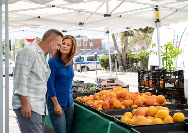 Imagem de um casal sorridente formado por um homem e uma mulher em frente a uma banca de frutas em uma feira de rua para ilustrar matéria sobre as feiras em SP