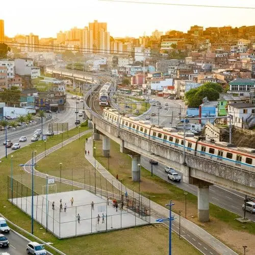 Imagem da paisagem urbana ao pôr do sol mostra o trem do metrô Salvador em um trilho elevado, carros nas ruas próximas e pessoas brincando em uma quadra cercada abaixo, com prédios ao fundo.