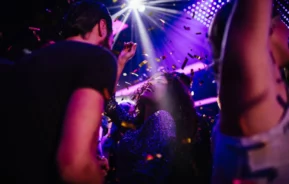 Imagem de pessoas dançando e comemorando sob luzes coloridas e confetes com um holofote brilhando acima em uma atmosfera festiva