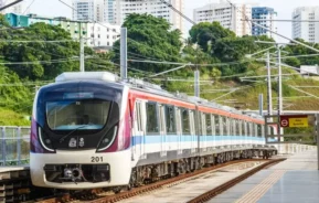 Imagem de um trem do metrô de Salvador estacionado em uma estação externa com trilhos visíveis e fios suspensos. Prédios altos e árvores verdes exuberantes surgem ao fundo sob um céu claro.