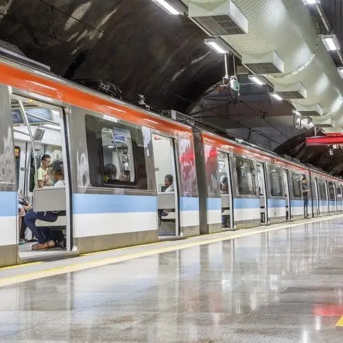 Imagem de um trem de metrô com listras azuis e brancas parado em uma estação do metrô em Salvador. Vários passageiros estão sentados em seu interior, enquanto a plataforma vazia é iluminada por uma forte luz no teto.