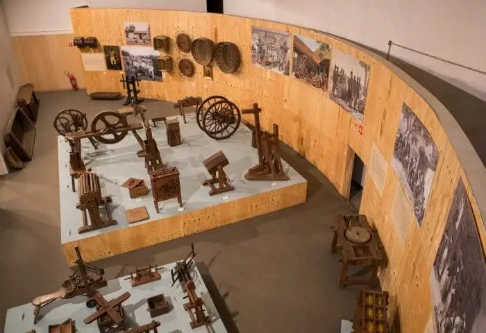 Sala de exposição do Museu Afro Brasil, no Parque do Ibirapuera, mostra ferramentas antigas de madeira, maquinário e artefatos em plataformas elevadas, com fotos históricas em preto e branco e cestos trançados montados em paredes de madeira clara.