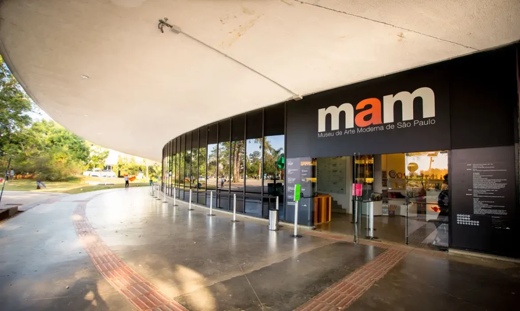 Imagem da entrada do Museu de Arte Moderna de São Paulo, com grandes janelas de vidro e arquitetura moderna. O acrônimo mam do museu é exibido acima da porta. As árvores e o parque sugerem atividades para quem procura o que fazer no Parque Ibirapuera.