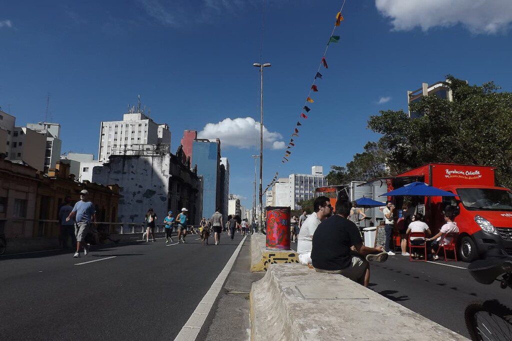 Pessoas sentadas no batente, no Minhocão. Há algumas pessoas caminhando e food trucks.