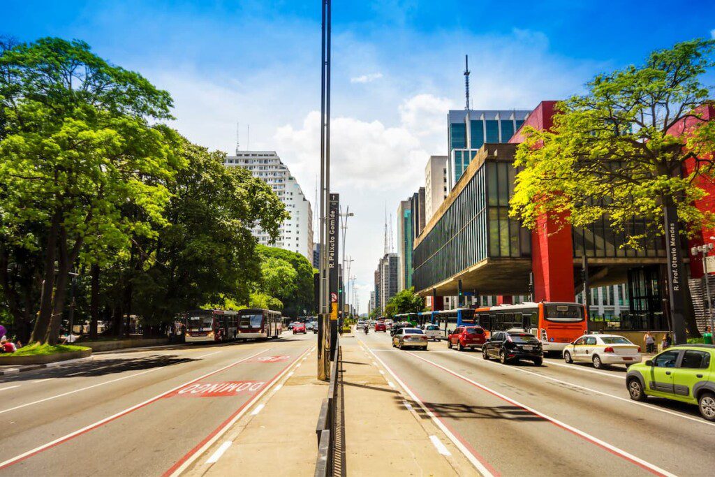 MASP, à direita da imagem, sustentado por colunas vermelhas. No centro da foto vemos uma avenida movimentada.