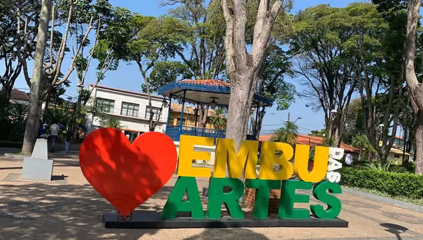 Imagem de uma grande placa colorida diz “Embu das Artes” com um coração vermelho na frente, em um parque ensolarado com árvores, um gazebo com telhado azul e prédios distantes