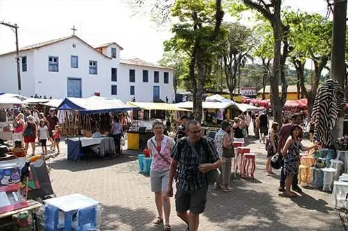 Imagem de pessoas caminhando entre barracas em Embu das Artes, com árvores fazendo sombra e um prédio branco com portas e janelas azuis ao fundo. Vários produtos são exibidos sob barracas.