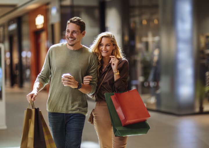 Imagem de um casal sorridente andando pelos corredores de um shopping com algumas sacolas de compras para ilustrar matéria sobre os shoppings no rj