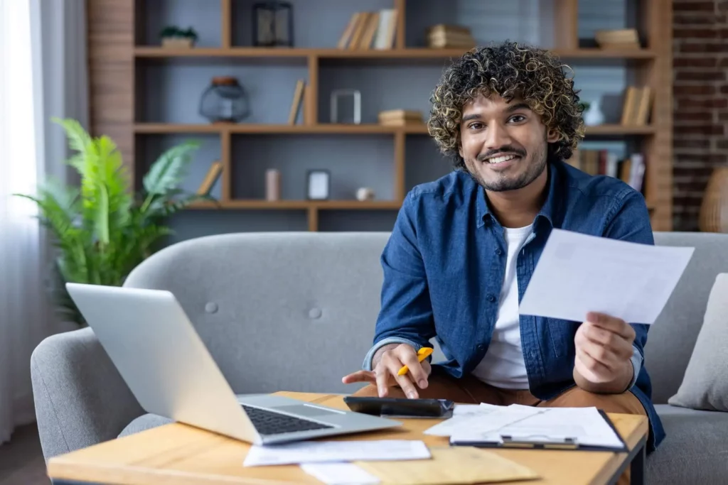 Imagem de um homem sorridente sentado em um sofá em uma mesa de centro com um laptop, calculadora e papéis em uma sala de estar aconchegante com prateleiras e plantas ao fundo