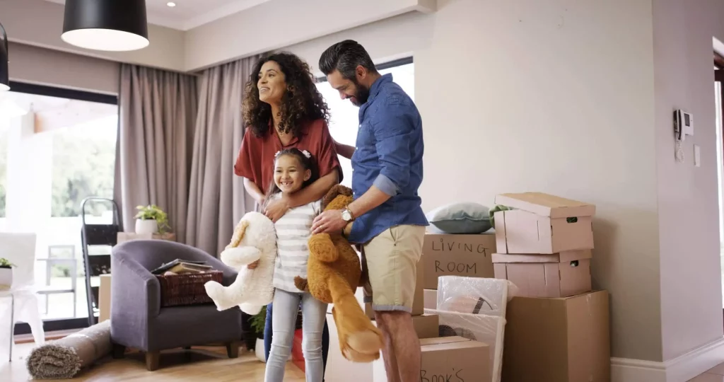 Imagem de uma família sorridente com uma menina está em uma sala de estar cercada por caixas de mudança, animada para se mudar para sua nova casa. A menina segura um bicho de pelúcia enquanto seus pais estão próximos, todos parecendo felizes