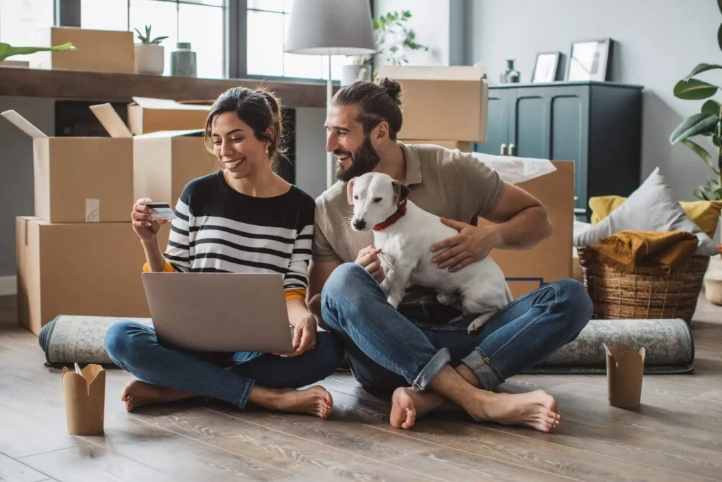 Imagem de um casal sorridente sentado no chão entre caixas de mudança, com um laptop e um cartão de crédito. O homem segura um cachorro branco no colo em seu quarto moderno e iluminado, cheio de itens embalados