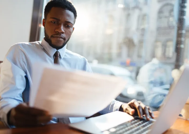 Um homem negro, vestido de roupa social, analisa uma documentação com auxílio do notebook.