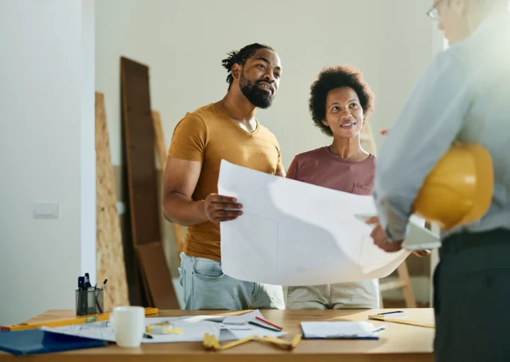 Imagem de um casal em uma residência em construção falando com um profissional da área para ilustrar matéria sobre área construída