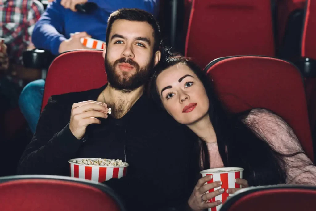 Imagem de um casal sentado junto em poltronas vermelhas de teatro, saboreando uma pipoca. A mulher encosta sua cabeça no ombro do homem enquanto assistem a um filme, cercados por outras pessoas ao fundo.