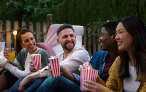 Imagem de quatro amigos sentados ao ar livre, sorrindo e conversando, cada um segurando um recipiente de pipoca listrado, para ilustrar matéria sobre os cinemas de rua em SP. Uma pessoa toma um drinque amarelo enquanto relaxam juntos, aproveitando a atmosfera casual com árvores e uma cerca de madeira atrás deles.