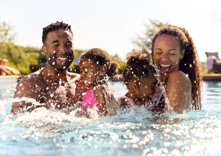 Imagem de uma família em uma piscina com duas meninas espirrando água para cima para ilustrar matéria sobre clubes em SP