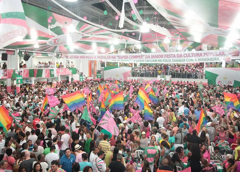 Quadra da escola de samba Mangueira, lotada de pessoas com bandeiras e blusas coloridas. 