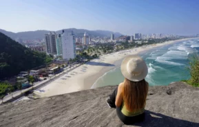 Imagem de uma mulher sentada em cima de um mirante no Guarujá, em São Paulo, observando a vista do mar, faixa de areia e prédios da cidade para ilustrar matéria sobre o que fazer no Guarujá