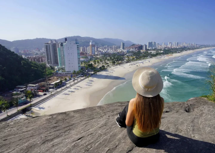 Imagem de uma mulher sentada em cima de um mirante no Guarujá, em São Paulo, observando a vista do mar, faixa de areia e prédios da cidade para ilustrar matéria sobre o que fazer no Guarujá