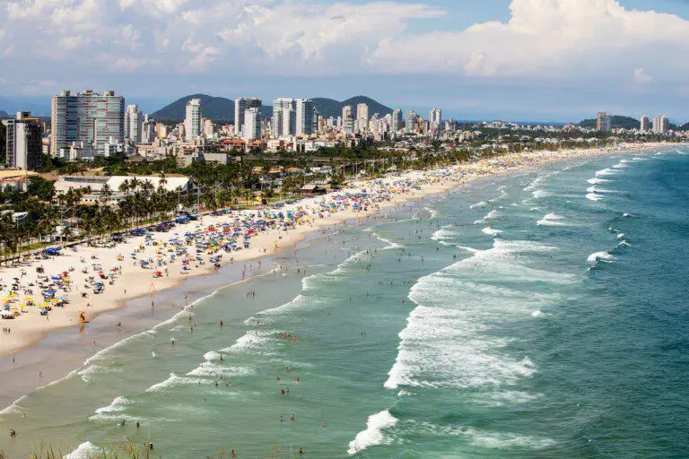 Vista para a Praia da Enseada, no Guarujá, mostra mar, faixa de areia, banhistas na praia, vegetação e prédios ao fundo