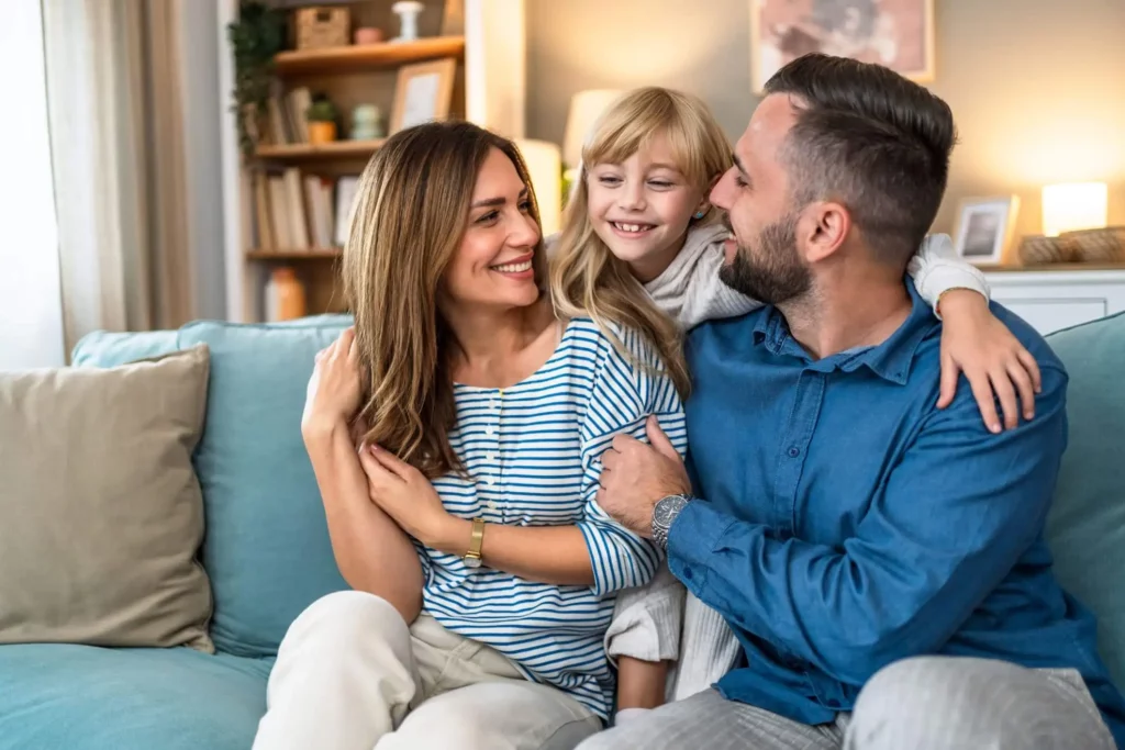 Imagem de uma família de três pessoas, incluindo uma mulher, um homem e uma menina, sentados em um sofá azul em sua aconchegante sala de estar
