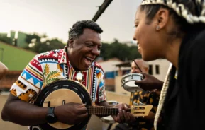 Imagem de um homem de camisa colorida sorrindo e tocando banjo, enquanto uma mulher de cabelos trançados ri ao lado. Outra pessoa toca pandeiro ao fundo em uma animada roda de samba no Rio de Janeiro, todos curtindo música ao ar livre juntos