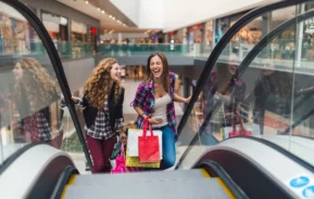 Imagem de duas mulheres jovens segurando sacolas em uma escada rolante de um shopping para ilustrar matéria sobre shopping em bh