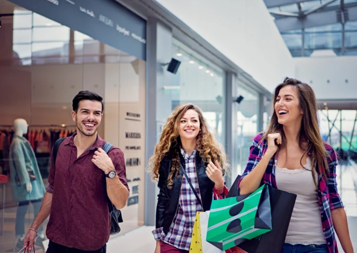 Imagem de três amigos, sendo duas mulheres e um homem, passeando sorridentes e com sacolas nas mãos pelo corredor de um shopping para ilustrar matéria sobre shopping em Goiânia