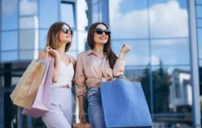 Imagem de duas mulheres jovens usando óculos escuros e sorrindo, segurando grandes sacolas de compras depois de fazer compras em um shopping ao ar livre em frente a um prédio de vidro em um dia ensolarado.