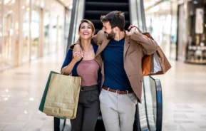 Imagem de um homem e uma mulher abraçados e felizes, descendo uma escada rolante com sacolas de compras nas mãos, para ilustrar matéria sobre shopping em Guarulhos