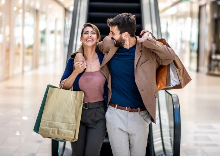 Imagem de um homem e uma mulher abraçados e felizes, descendo uma escada rolante com sacolas de compras nas mãos, para ilustrar matéria sobre shopping em Guarulhos