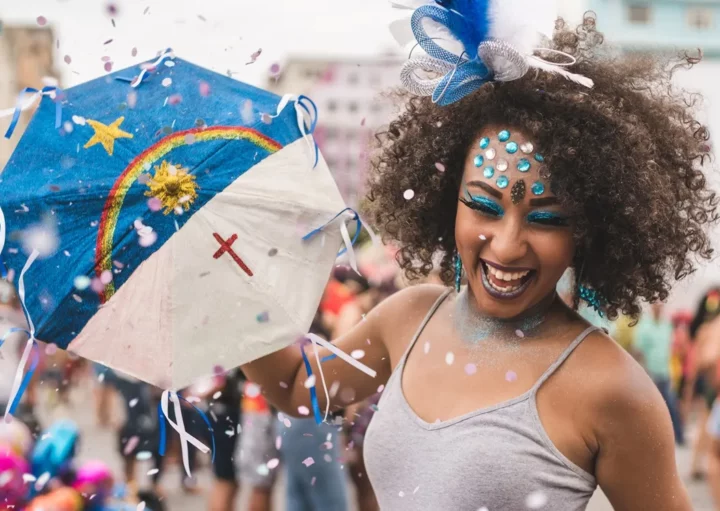 Foto que ilustra matéria sobre Carnaval de Recife mostra uma mulher negra, muito sorridente e enfeitada com glitter e brilhantes diversos se divertindo com uma pequena sombrinha usada para dançar frevo com estampa da bandeira do estado de Pernambuco. (Foto: Getty Images)