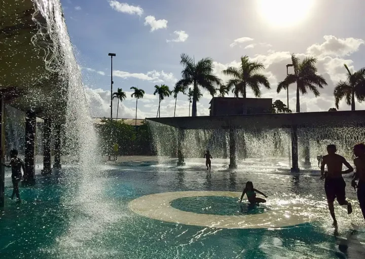 Foto que ilustra matéria sobre o Parque Madureira mostra crianças brincando em uma fonte artificial de águas ao cair do sol