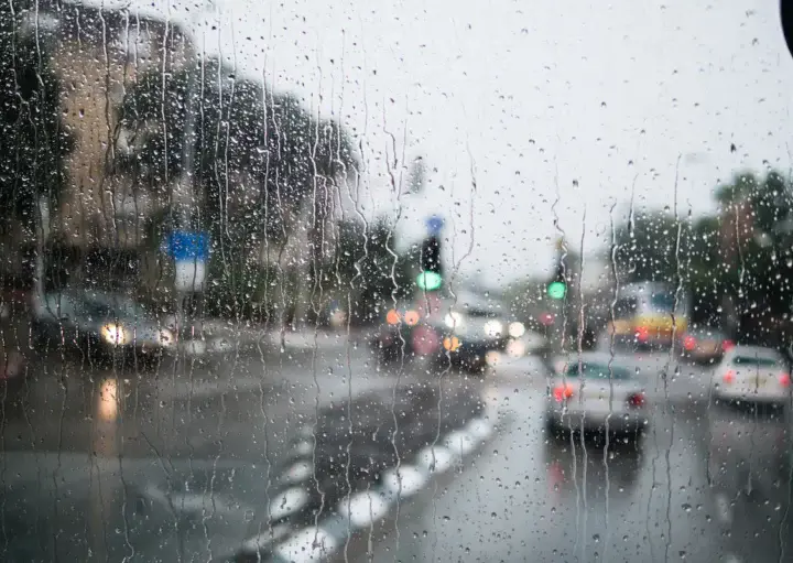  Imagem de um vidro embaçado e molhado pela chuva onde é possível ver a rua, carros e um semáforo aberto para ilustrar matéria sobre lugares para ir em dias de chuva no RJ