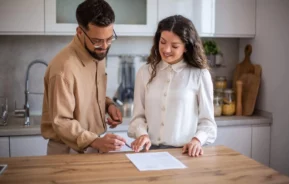 Imagem de um homem e uma mulher em uma cozinha, sorrindo e analisando um documento juntos em um balcão de madeira. O homem está segurando uma caneta, pronto para assinar, enquanto a mulher aponta para o papel
